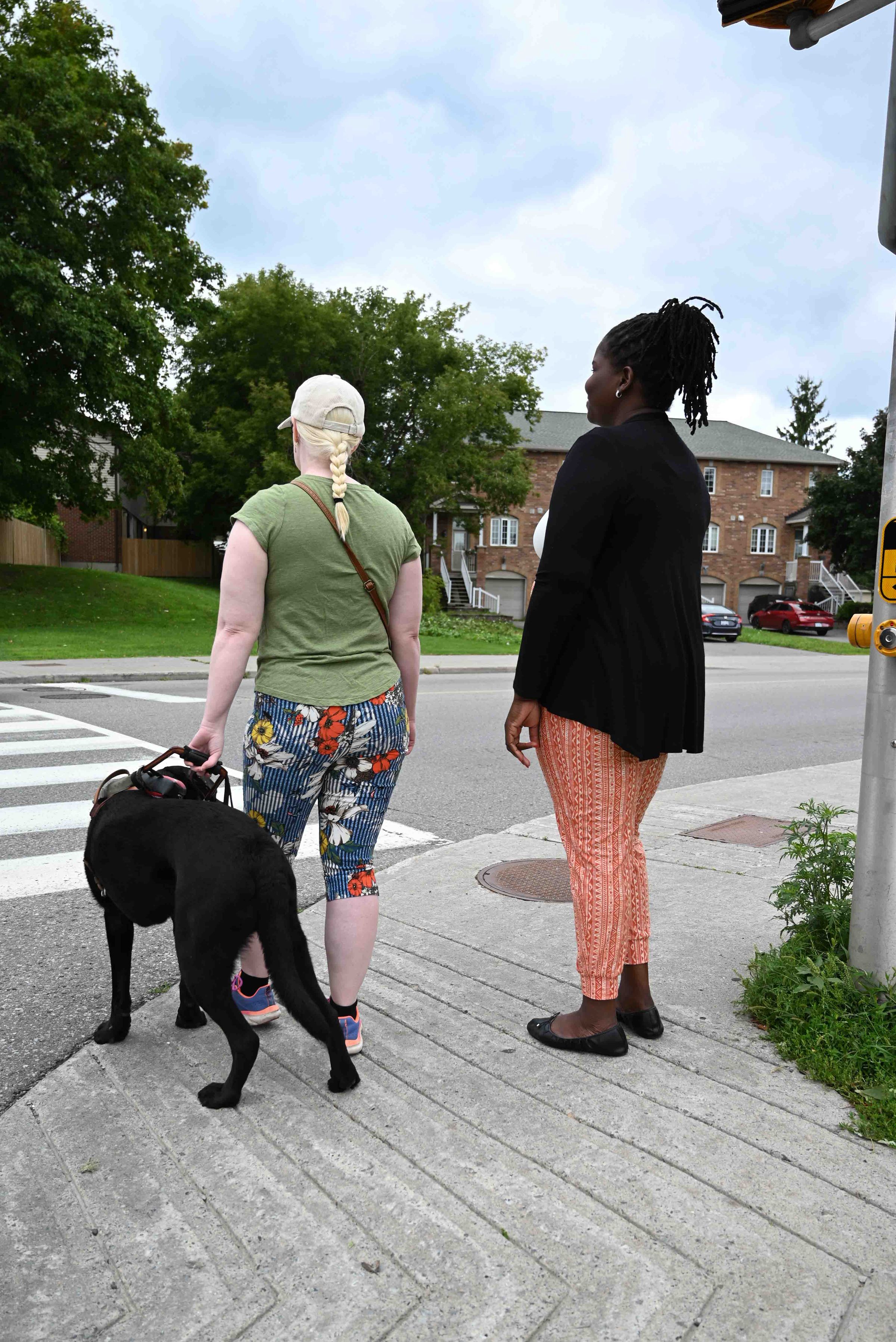 The image is a woman at an intersection corner facing the crosswalk with her guide dog. A second woman is standing beside her on the right slightly behind. 