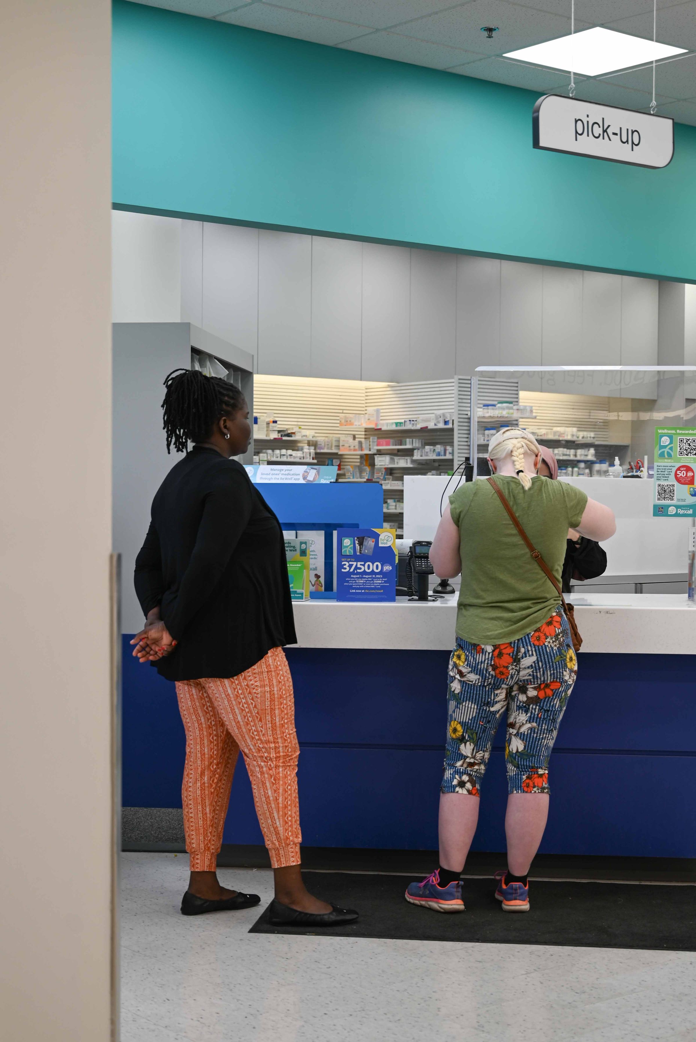 The image is of a woman standing at a pharmacy counter. There are two other people standing behind her, one of whom is across the counter. The pharmacy counter has a sign that says “pick up” above. The walls are blue and there are several shelves filled with items across the counter. The woman has a bag slung over her shoulder and her head held forward focusing on something she is holding. Only her back is visible.