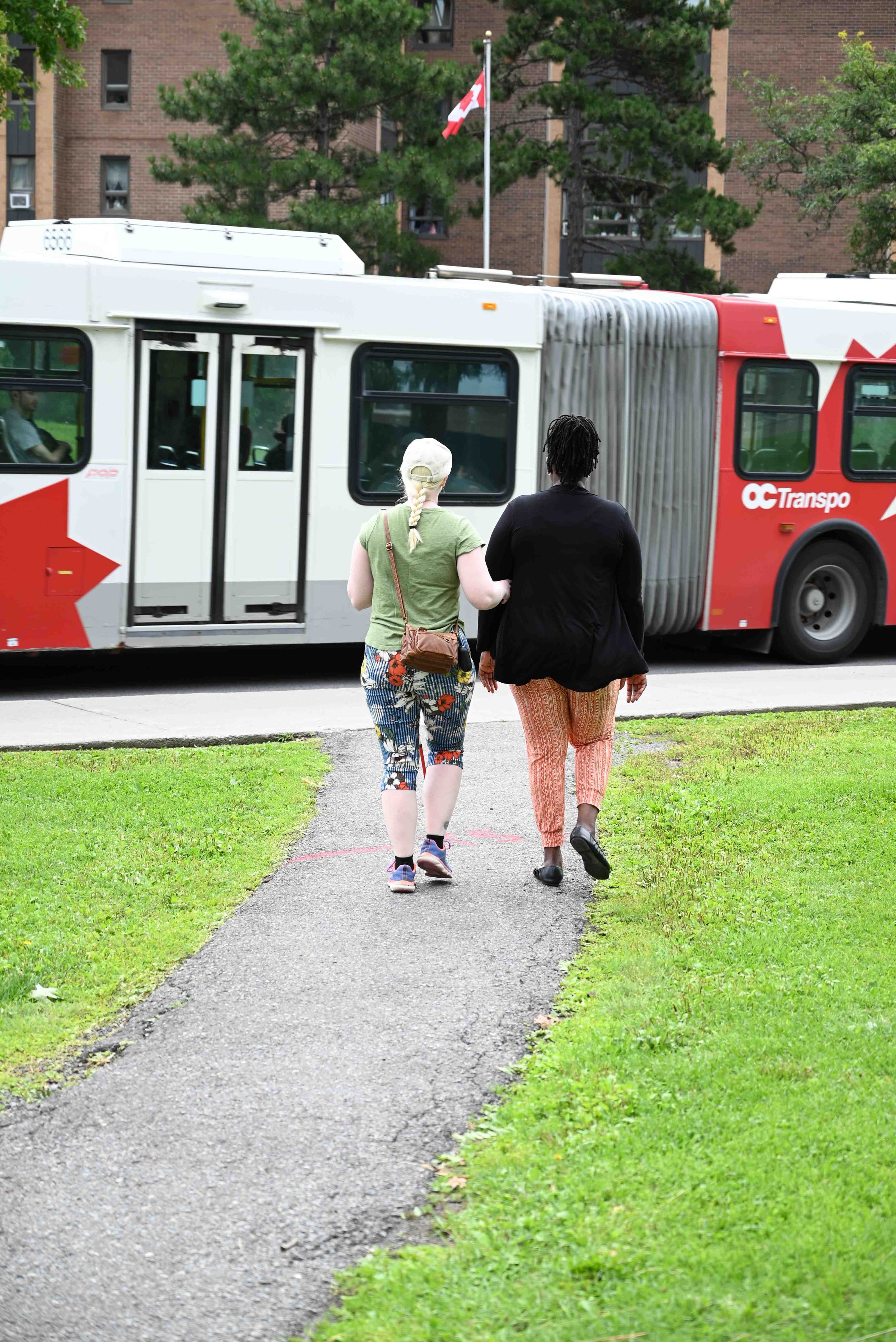 This image showcases two women walking together on a sidewalk, using the standard human guide grip. They are heading towards a city transit bus labelled "OC Transpo," which is parked by the side of the road.